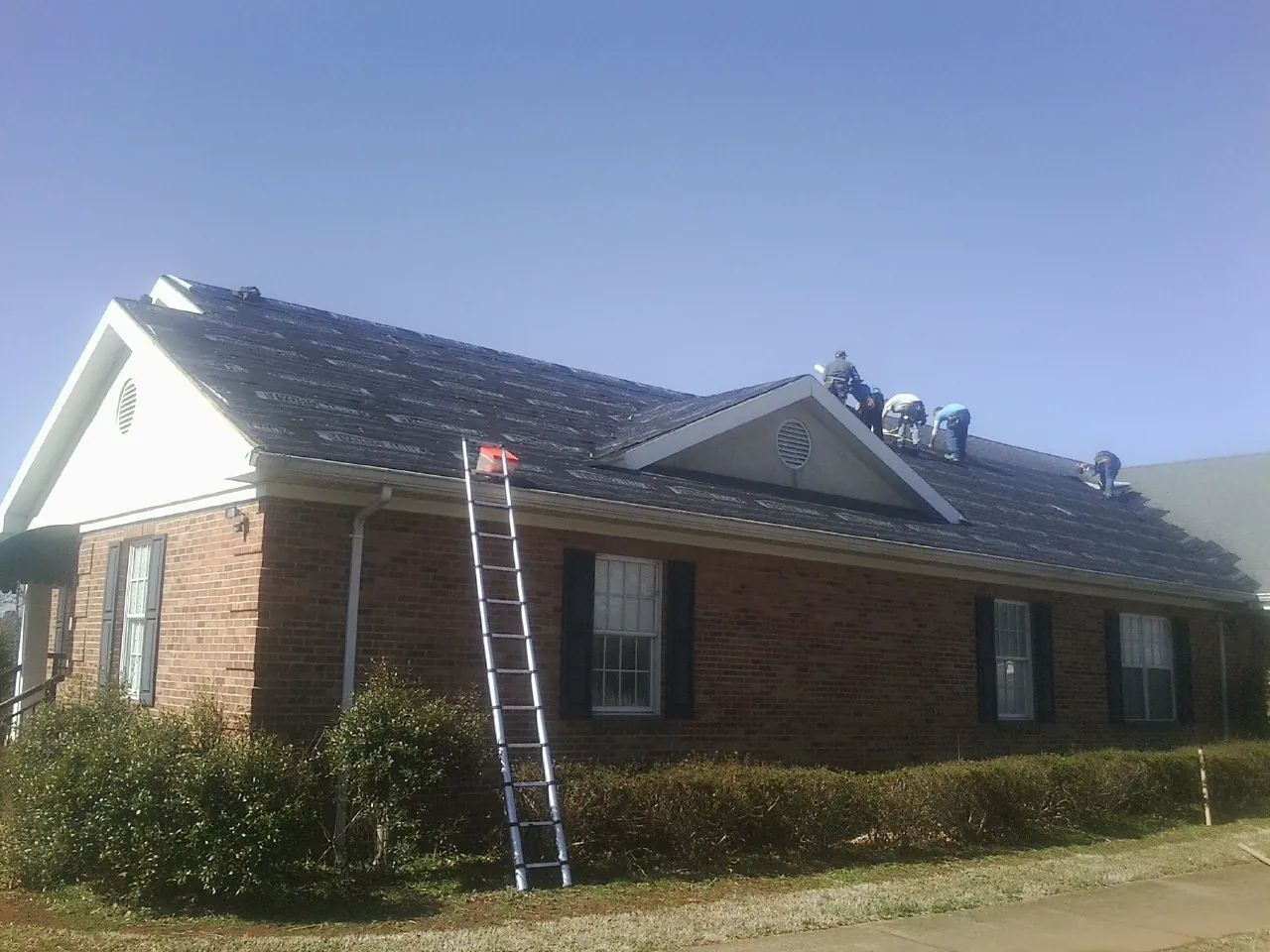 Crew tearing shingles off a brick ranch roof with a ladder set — Emergency Roof Repair work by our crew in Bennett