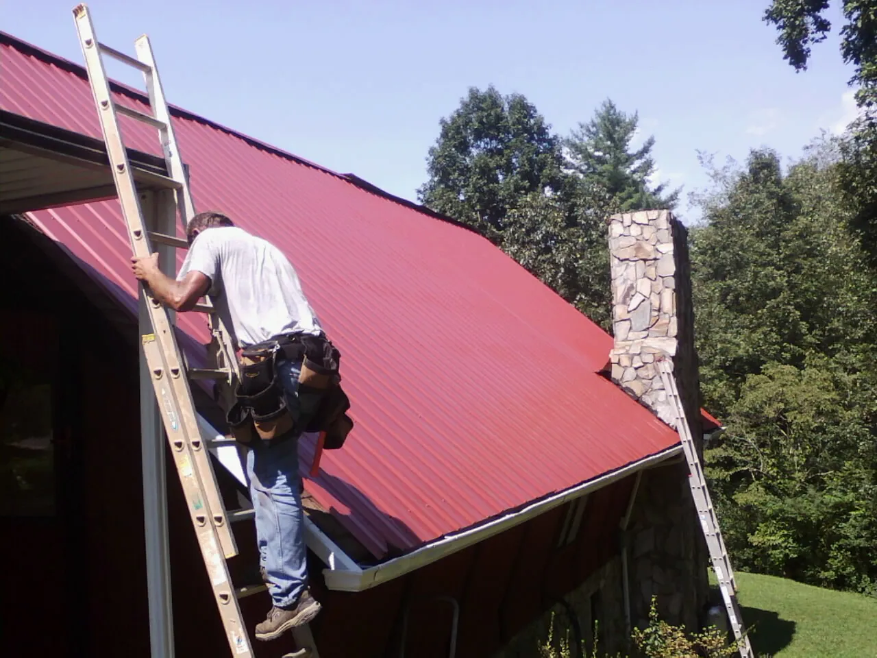 Roofer on a ladder beside a red metal roof and stone chimney — Soffit & Fascia Repair by Roofing Brothers in Bennett
